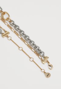 Quattro braccialetti in catena strati d'oro e argento con vari collegamenti e chiusure, uno pendente inciso con "DKNY" su uno sfondo chiaro.