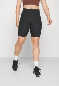 Černé cyklistické kraťasy s vysokým pasem, vyrobené z elastického materiálu, s bočními švy a hladkou texturou, doplněné černými sportovními botami a bílými ponožkami.