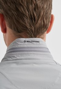 Hellgrauer Jackenkragen mit strukturierter Oberfläche, einem grauen Reißverschluss und dem in Schwarz gestickten Markennamen "MILESTONE".