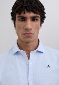 Cămașă cu dungi albastru deschis, cu guler bughea, logo mic cu craniu și oase încrucișate, și textură moale a țesăturii.