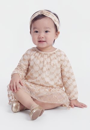 Petite fille assise avec une robe rose clair et un bandeau assorti avec des motifs papillons dorés et des chaussures dorées sur fond blanc.