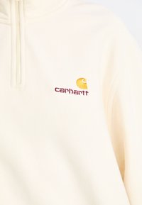 Pullover in pile crema con zip a un quarto. Presenta un logo Carhartt ricamato in giallo e bordeaux sul lato sinistro del petto. Texture morbida.