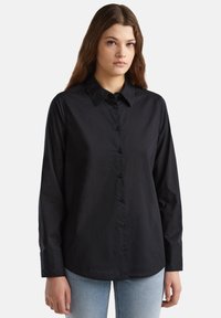 Camicia nera con colletto, maniche lunghe e texture liscia. Presenta bottoni neri e un orlo curvo, abbinata a jeans azzurri chiari.