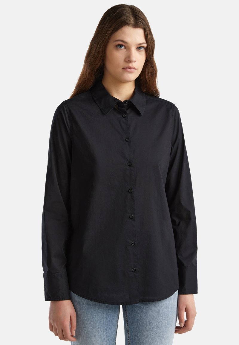 Camicia nera con colletto, maniche lunghe e texture liscia. Presenta bottoni neri e un orlo curvo, abbinata a jeans azzurri chiari.