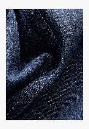 Tamno plavi traper od tkanine sa blagim sjajem, prikazujući mekan i naboran teksturu te vidljive šavove u kontrastnoj bež pređi.