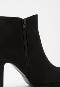 Bottine noire en faux daim avec un talon haut, dotée d'une fermeture éclair latérale et d'une texture lisse. Design épuré sans motifs ni ornements visibles.