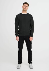 Svart långärmad sweatshirt med rund halsringning, ribbade manschetter och logotypdetalj vid fållen, kombinerad med svarta jeans och vita sneakers.