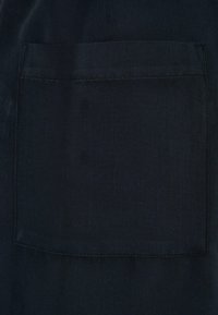 Tissu noir présentant une poche plaquée rectangulaire avec des coutures visibles. La texture semble lisse avec un léger éclat.