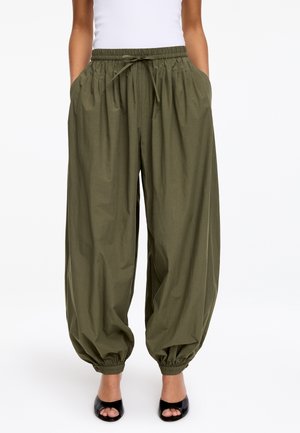 Kobieta w oliwkowozielonych, luźnych spodniach ze ściągaczami przy nogawkach, pasem na sznurek i bocznymi kieszeniami, w połączeniu z czarnymi sandałami na otwartym palcu.
