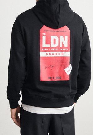 Personne portant un sweat à capuche noir avec un grand graphisme rouge et blanc dans le dos affichant "TO LDN," noms de villes, "FRAGILE," et "31 KG" écrit à la main.