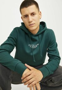 Grüne Baumwollkapuzenjacke mit Kordelzügen, Fronttasche und "JACK & JONES PREMIUM"-Logo. Das Modell posiert mit gefalteten Händen.