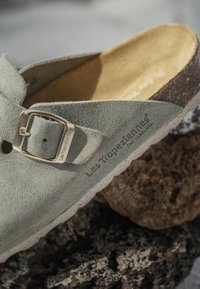 Zoccolo in suede grigio con un ampio cinturino, fibbia argentata e soletta in pelle beige. Presenta una finitura testurizzata e una suola in gomma di colore chiaro.
