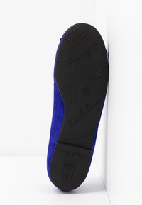 Semelle d'une chaussure plate bleue montrant une semelle extérieure en caoutchouc noir avec de petits motifs de chaussures de ballet en relief et nom de la marque "PrettyBallerinas."