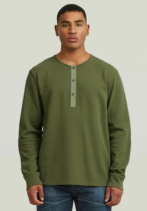 Unge mand iført olivengrøn langærmet wafflestrikket henley-shirt med tre knapper og blå jeans, stående mod en ensfarvet baggrund.