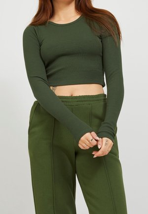 Woman wearing a fitted, long-sleeve dark green crop top and loose dark green pants, with visible tattoos on her left wrist and side.