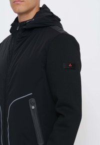 Uomo con giacca nera con cappuccio, chiusura frontale a cerniera e tasca con cerniera sul lato sinistro, logo visibile sulla manica sinistra.
