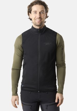 Black vest with a zip closure and a fitted design, paired with a long-sleeve olive green top. Features a logo and zippered arm pocket.