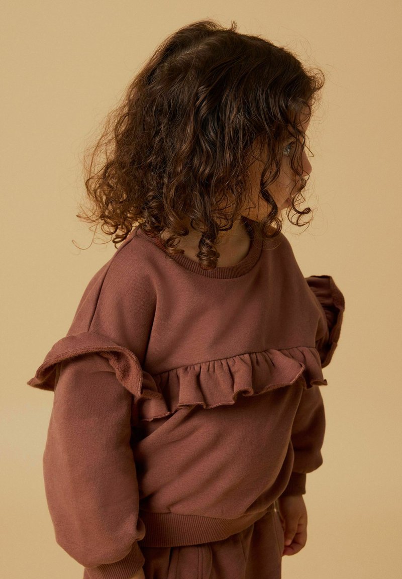 Jeune enfant aux cheveux bouclés et foncés portant un sweat-shirt marron à volants, regardant sur le côté sur un fond beige.