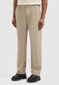 Beige Hosen mit lockerer Passform, ausgestattet mit einem Knopf und einem elastischen Bund, aus glattem Stoff. Kombiniert mit schwarzen Schuhen.