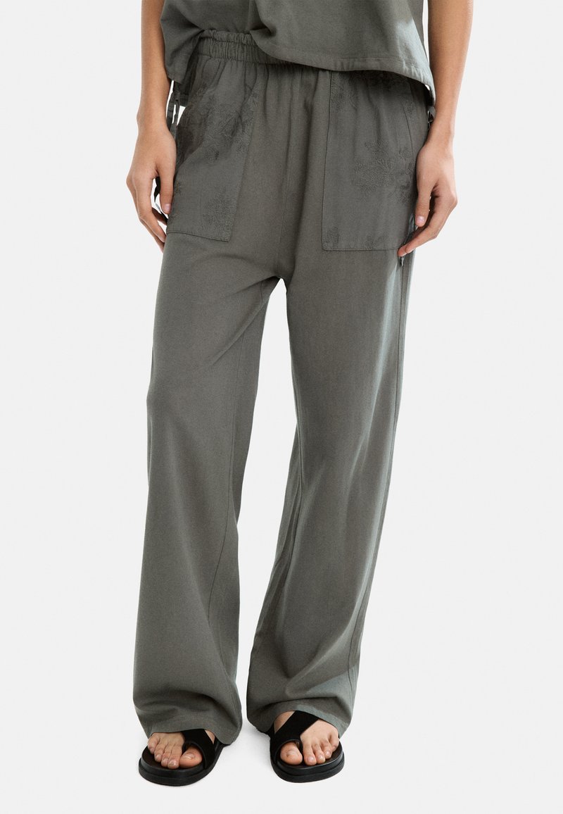 Szerokie spodnie w miękkiej, szarej tkaninie z elastycznym pasem, z dwoma przednimi kieszeniami i minimalnymi akcentami teksturowymi. W zestawieniu z czarnymi sandałami.
