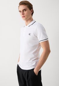 Bílá polo košile s krátkým rukávem a černými lemy na límci a rukávech. Na levé části hrudi je vyšitá nášivka "CK". Materiál je bavlněný.