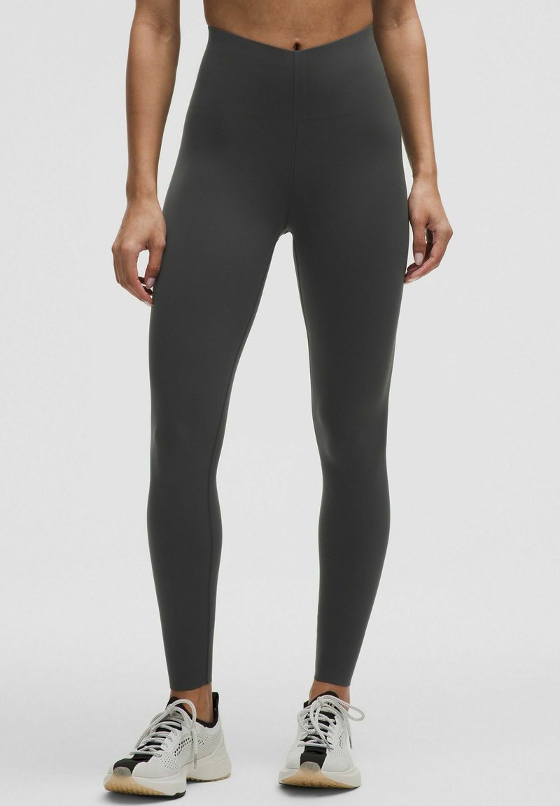 Leggings a vita alta, aderenti, in materiale grigio scuro con una texture liscia, abbinati a scarpe sportive leggere con accenti neri.