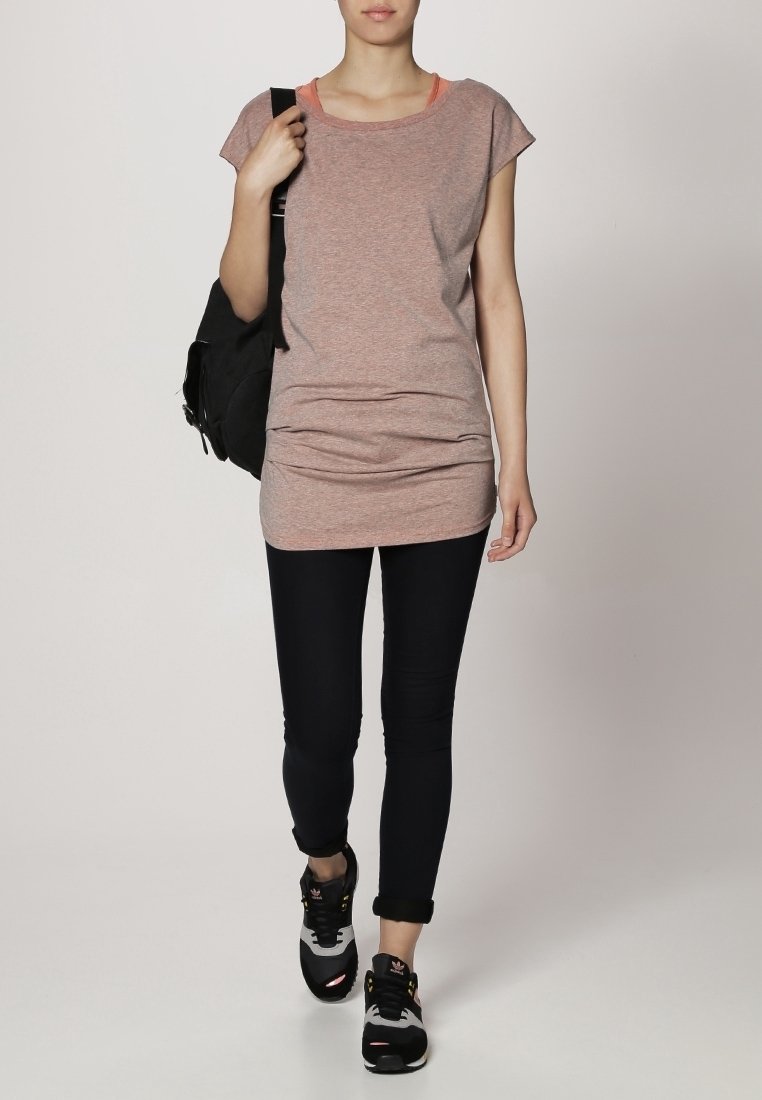 T-shirt gris à manches courtes et un dos légèrement plus long, associé à un jean skinny noir et des chaussures de sport noires. Sac à dos noir porté sur l'épaule.