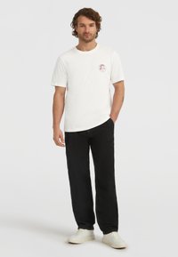 T-shirt bianca in cotone con una piccola stampa grafica rossa, abbinata a pantaloni neri larghi e sneakers bianche. Design semplice, stile casual.