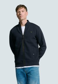 Marineblauwe zip-up jas met een textuurpatroon, staande kraag en gouden rits. Voorzien van zijzakken en een subtiele logo-detail op de borst.