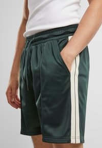 Shorts de sport vert foncé en matériau lisse, dotés d'une taille avec cordon de serrage et de rayures blanches sur les côtés pour un contraste. Conçus avec des poches latérales.