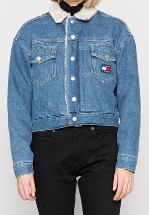 Person iført en blå denimjakke med hvidt imiteret pelskrave og knaplukning, kombineret med sorte bukser.