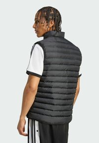 Ung mand med fletninger iført en sort puffervest over en hvid t-shirt med korte ærmer og sorte og hvide stribede bukser, vender mod venstre.
