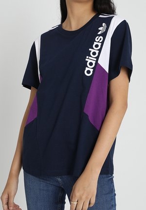 T-shirt Adidas bleu marine avec des panneaux blancs et violets, porté par une personne, avec le logo Adidas et du texte en haut à gauche sur le devant.