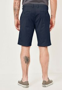 Marineblau Shorts aus einem glatten Stoff, mit zwei Gesäßtaschen und einem slim fit Design. Das Modell trägt graue Sneakers.