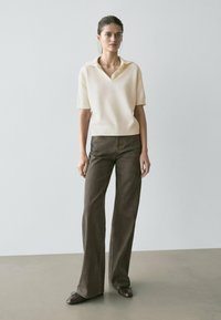 Cremefarbener kurzärmeliger Polo-Pullover mit V-Ausschnitt, kombiniert mit hoch taillierter, dunkelbrauner Schlaghose und braunen Slippern.