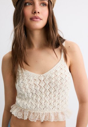 Giovane donna con lunghi capelli castani che indossa un top corto di maglia color crema con bordo in pizzo e un cappello intrecciato beige, su uno sfondo chiaro uniforme.