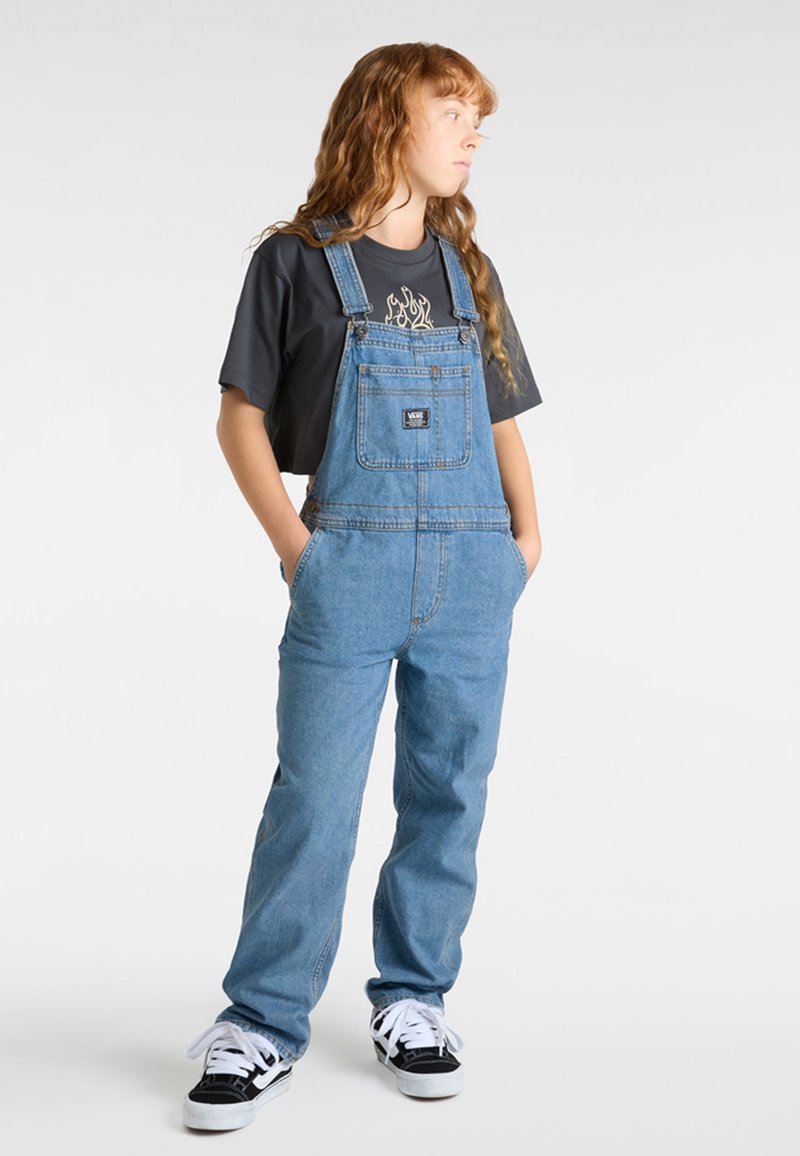 Peto de denim en azul claro con tirantes ajustables, bolsillo frontal y corte relajado; combinado con una camiseta gráfica de manga corta negra y zapatillas.