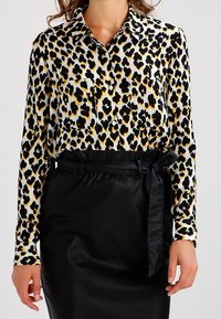 Blusa de estampado de leopardo en blanco, negro y dorado, con cuello y dos bolsillos en el pecho, combinada con una falda de satén negra con cinturón.