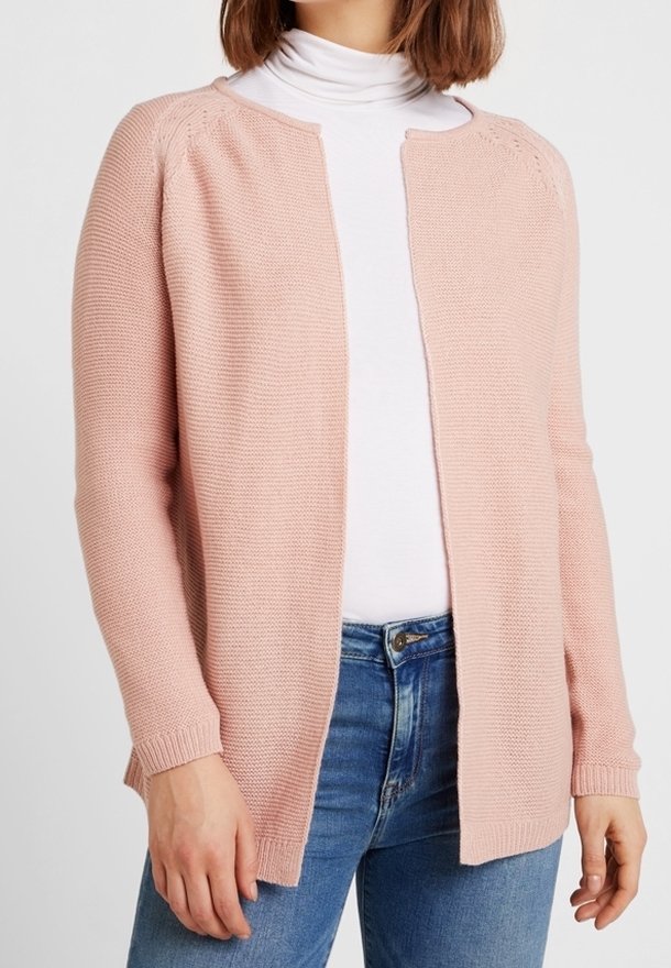 Femme portant un cardigan rose clair en maille à ouverture frontale, un col roulé blanc et un jean bleu, vue des épaules aux hanches.