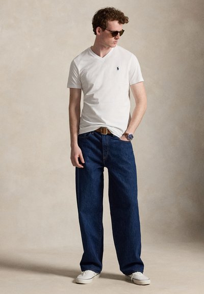 Muž so slnečnými okuliarmi, bielym tričkom s výstrihom do V, modrými širokými džínsami, hnedým opaskom, náramkovými hodinkami a bielymi teniskami, stojaci pred neutrálnym pozadím.