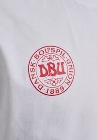 Rood rond logo met "DBU" in het midden, omgeven door "Dansk Boldspil-Union 1889" op een witte stoffen achtergrond.