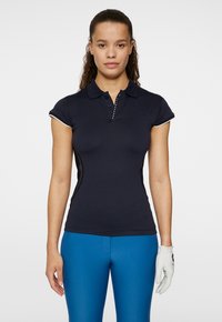 Marinblå polo med korta ärmar och knäppning, med vita detaljer och dekorativa nitar. Matchas med blåa tajta byxor och en vit handske.