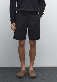 Zwarte katoenen shorts met een taille met trekkoord, gladde textuur, knielengte, gecombineerd met bruin instap schoenen; tegen een neutrale achtergrond.
