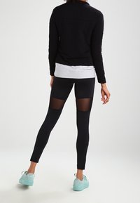 Sorte atletiske leggings med gennemsigtige mesh-paneler på de øverste lår, parret med en grå top og en sort jakke samt mintgrønne sneakers.