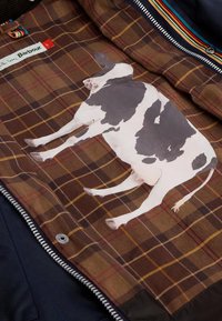 Η γραμμή της καφέ καρό ζακέτας διαθέτει μια μεγάλη εικονογράφηση μιας μαύρης και άσπρης αγελάδας, με μαλακή υφή και ζωντανά ραψίματα κοντά στο φερμουάρ.