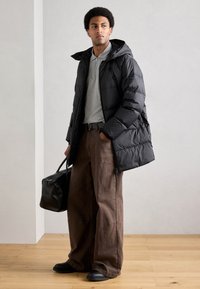 Casaco acolchoado preto com capuz, camisa polo cinza, calças largas castanhas e mala preta texturizada. Calçado preto completa o conjunto.