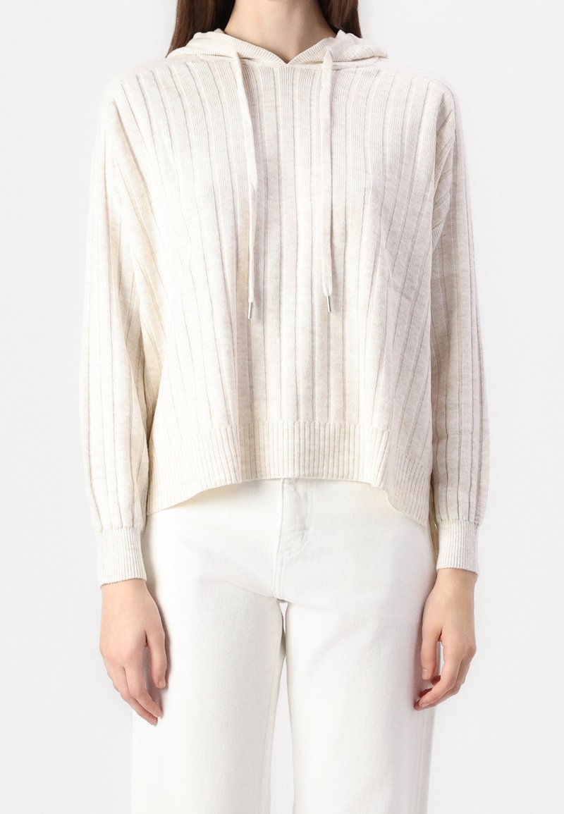 Femme portant un sweat à capuche en maille côtelée crème avec cordons et un pantalon blanc taille haute, debout devant un fond uni.