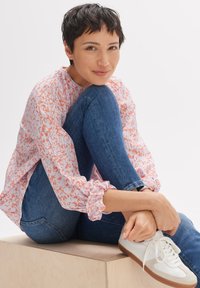 Blusa rosa chiaro con macchie arancioni, tessuto testurizzato, maniche a polsino, abbinata a jeans di denim blu e sneakers bianche su una piattaforma di legno.