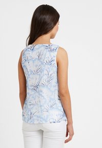 Mujer con cabello largo y oscuro, vestida con una blusa sin mangas de flores azules y blancas y pantalones blancos, vista desde atrás contra un fondo blanco.