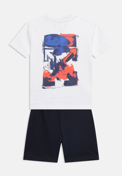 Λευκό πουκάμισο με κοντά μανίκια και αφηρημένα μπλε και κόκκινα γραφικά στην πλάτη, σε συνδυασμό με σκέτα μαύρα σορτς, εμφανίζεται σε λευκό φόντο.